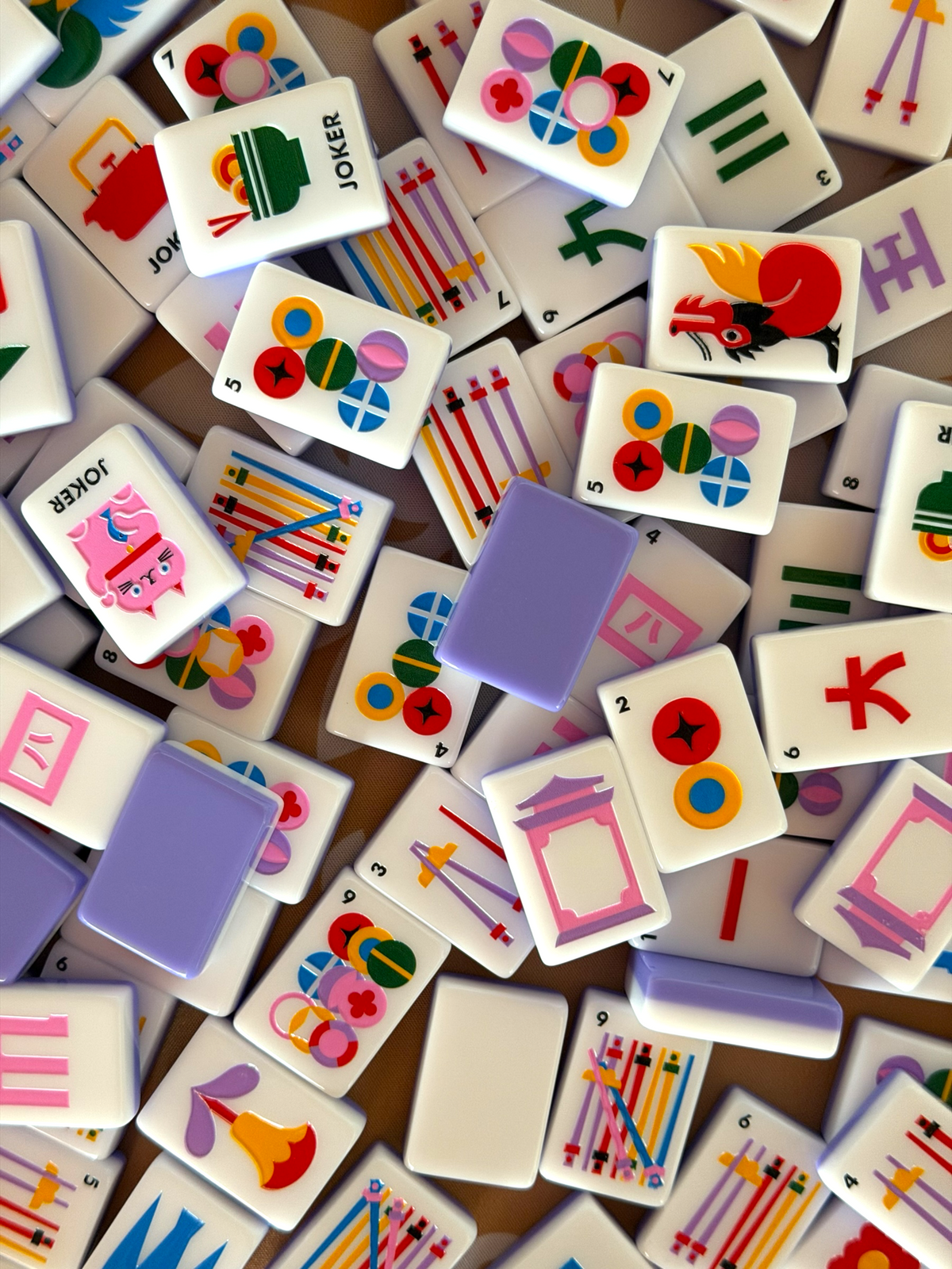 A close-up, top-down view of a mahjong tile set scattered across a surface. The tiles are primarily white with various colorful printed symbols, characters, and illustrations, including jokers and traditional Chinese-style motifs.